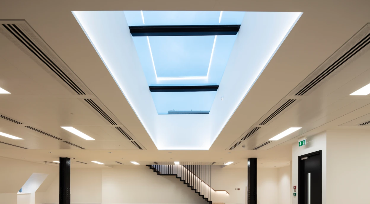 A modern, spacious room with light hardwood flooring and minimalist design. A large skylight in the ceiling allows natural light to enter, and a sleek staircase is visible in the background. Walls are painted white, enhancing the bright and airy atmosphere.