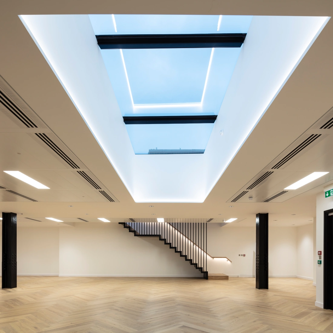 Intérieur moderne avec un plafond lumineux et un puits de lumière. Un escalier en bois blanc mène à un niveau supérieur. Le sol est en parquet clair et les murs sont peints en blanc.