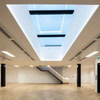 A modern, spacious room with light hardwood flooring and minimalist design. A large skylight in the ceiling allows natural light to enter, and a sleek staircase is visible in the background. Walls are painted white, enhancing the bright and airy atmosphere.