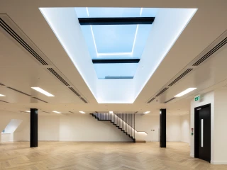 Intérieur moderne avec un plafond lumineux et un puits de lumière. Un escalier en bois blanc mène à un niveau supérieur. Le sol est en parquet clair et les murs sont peints en blanc.