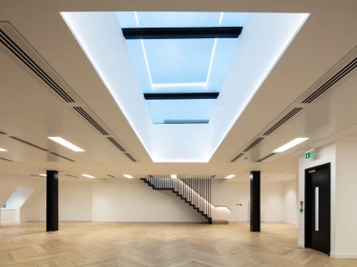 Offener Raum mit hellen Wänden und einem großen Dachfenster. Dezente LED-Beleuchtung und elegante Holztreppe im Hintergrund. Der Boden ist aus Holz mit einem Fischgrätenmuster.