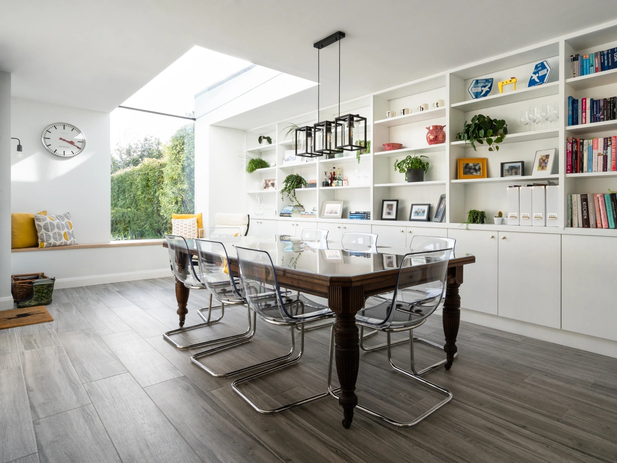 Comedor moderno con una mesa de madera y sillas transparentes. Estanterías blancas llenas de libros y decoraciones. Gran ventana que permite la entrada de luz natural.
