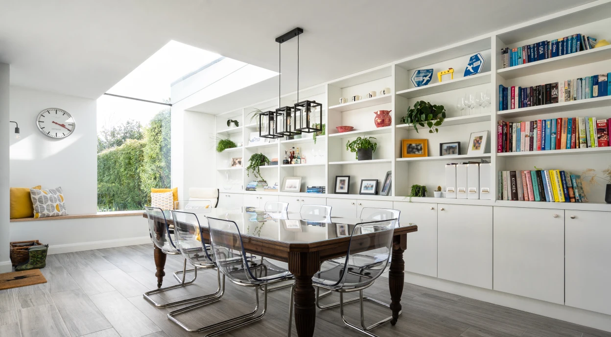 A modern dining area featuring a large wooden table surrounded by transparent chairs. There's a sleek light fixture hanging above the table. The walls have built-in shelves displaying books and decorative items, with a large window letting in natural light. A cozy seating nook with pillows is visible in the background.