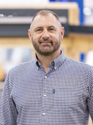 A man with a beard and short hair stands smiling in a workshop setting. He is wearing a checkered shirt and there are wooden structures and equipment in the background.