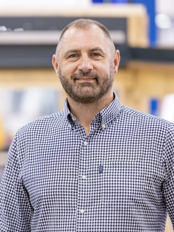 A man with a beard and short hair stands smiling in a workshop setting. He is wearing a checkered shirt and there are wooden structures and equipment in the background.