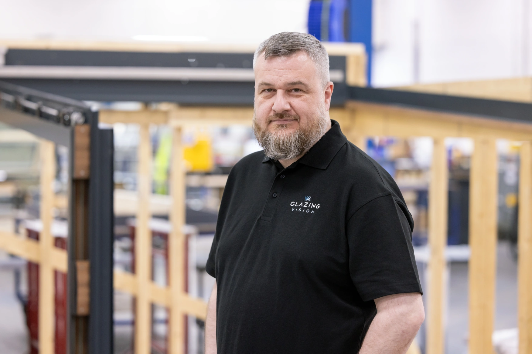 A man with a beard poses confidently in a black polo shirt, standing in a workshop filled with wooden frames and machinery.