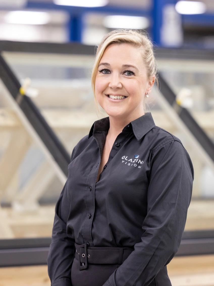 A woman in a black shirt stands smiling in front of large windows in a workshop. The background shows wooden frames and a well-lit setting.