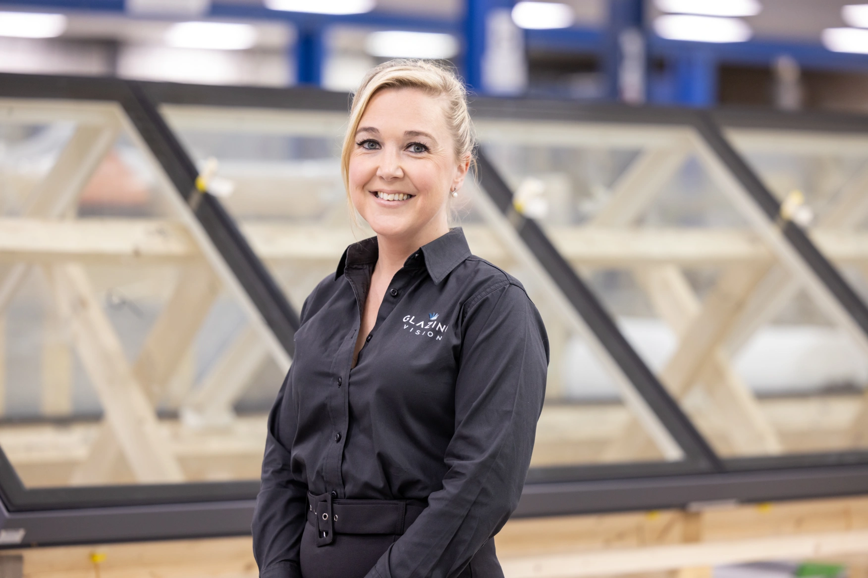 A woman with blonde hair styled in a ponytail smiles while standing in front of large windows at a manufacturing facility. She is wearing a black shirt with a logo on it. The background shows a well-lit workspace filled with window frames.