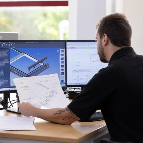 A person sitting at a desk, reviewing engineering plans while looking at a computer screen displaying a 3D model. The workspace includes multiple monitors, paperwork, and a bright background with windows.