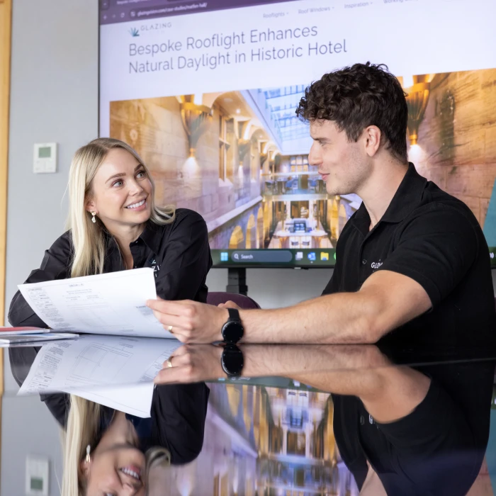 Two people are engaged in a discussion at a conference table, reviewing plans and documents. A large display in the background features an article about enhancing natural daylight in a historic hotel. Both individuals are wearing black shirts with the company's logo.