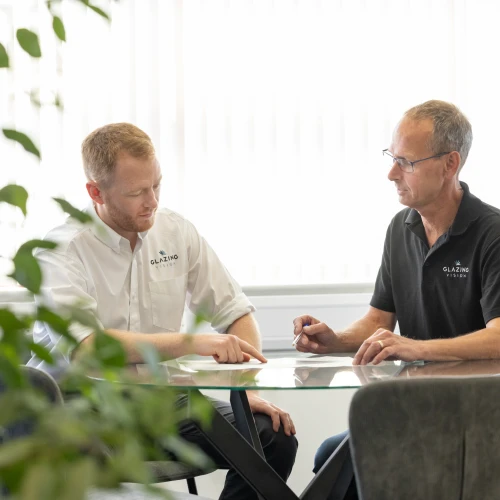 Two men engaged in a discussion at a glass table in a modern office setting. One man points at a document while the other takes notes. Green foliage is visible in the foreground.