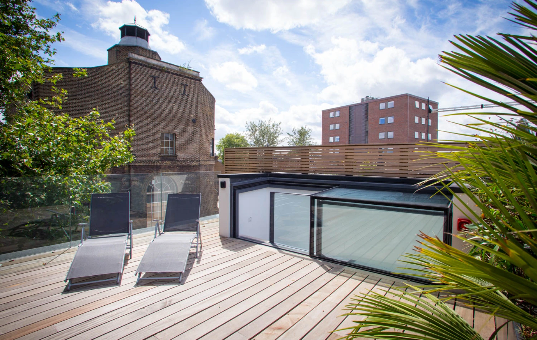 A rooftop terrace features two lounge chairs on wooden decking, surrounded by greenery. In the background, a historic brick building and a modern apartment complex are visible under a partly cloudy sky.