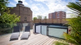 A spacious wooden deck features two lounge chairs under a bright sky with scattered clouds. In the background, a historic brick building and a modern apartment building are visible. Lush green foliage surrounds the area, creating a relaxing atmosphere.