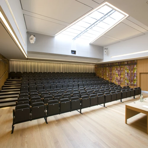 A modern auditorium featuring a tiered seating arrangement with black chairs, wooden panels, and a large skylight. A wooden table with chairs is positioned in the front near a podium. The walls include a colorful abstract design.