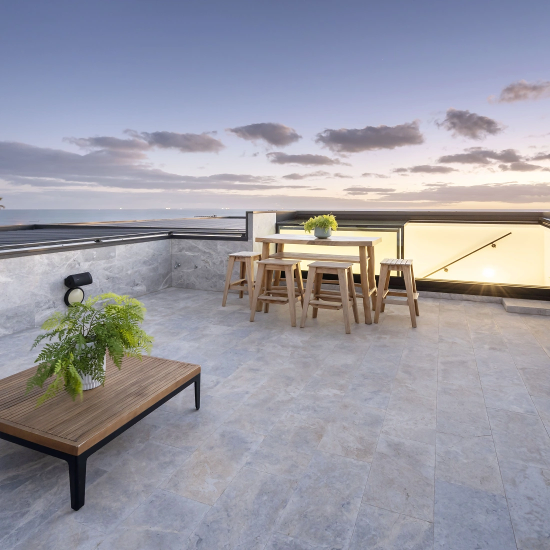 A rooftop terrace features a wooden outdoor table with four stools, a small coffee table, and a potted fern. The background shows a beautiful sunset with clouds and hints of palm trees.