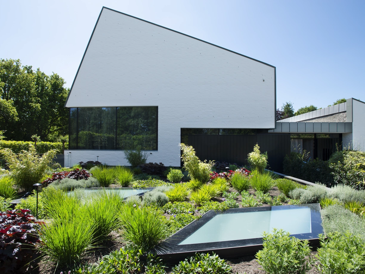 Une maison moderne à l’architecture angulaire, avec des murs blancs et de grandes fenêtres. Le jardin est verdoyant, rempli de plantes, d’herbes et de bassins d’eau rectangulaires. Le ciel est dégagé et ensoleillé.