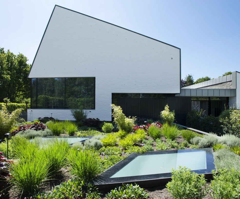 A modern white house with a distinctive angular roof is surrounded by lush greenery and landscaped gardens. Two shallow water features are integrated into the garden, reflecting the vibrant foliage. Bright blue sky is visible above, enhancing the serene outdoor atmosphere.