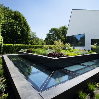 A modern building with a white facade sits beside a landscaped garden featuring a geometric arrangement of plants and glass features. The garden has varying greenery and colorful flowers, and the building boasts large windows reflecting the bright blue sky.