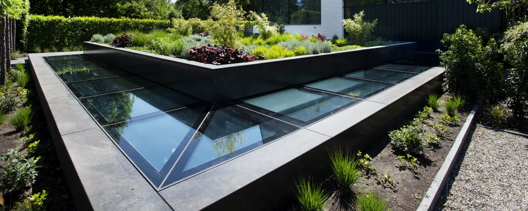 Maison moderne à l’architecture angulaire avec un jardin paysager en terrasse. Des plantes variées et des éléments en verre sont intégrés au design extérieur. L’éclairage naturel met en valeur la verdure environnante.