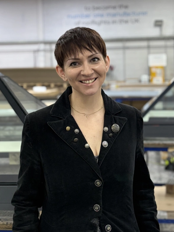 A woman with short, dark hair smiles confidently while wearing a black suit jacket. She stands in an industrial setting with glass structures and equipment in the background.