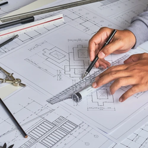 A person's hands are positioned over architectural blueprints while using a ruler and pencil. Various drafting tools, including a compass and pencils, are scattered on the table surrounded by rolled-up paper sheets.