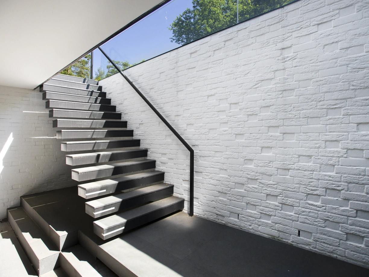Modern staircase leading upwards, featuring sleek wooden steps and a glass railing. The walls are textured and white, with natural light coming in from above, illuminating the space.