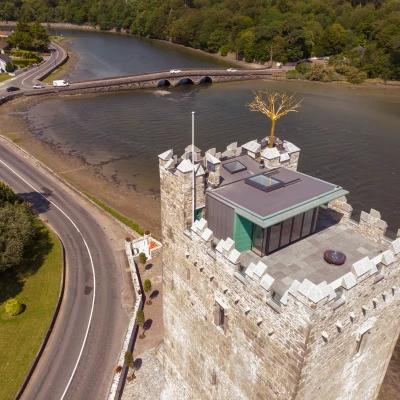 Aerial view of a stone tower with a modern glass structure on top, located near a river. Surrounding greenery and a road alongside the water are visible. A bridge crosses the river in the background.