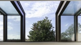 Large windows with a modern design open up to a view of a green tree and blue sky with scattered clouds. The interior features light-colored walls, wooden flooring, and minimalist furniture.