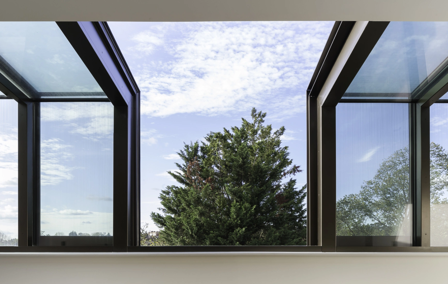 Ventana amplia con marcos oscuros, mostrando un paisaje exterior con un árbol y cielo parcialmente nublado. Interior moderno con pared blanca y suelos de madera clara.