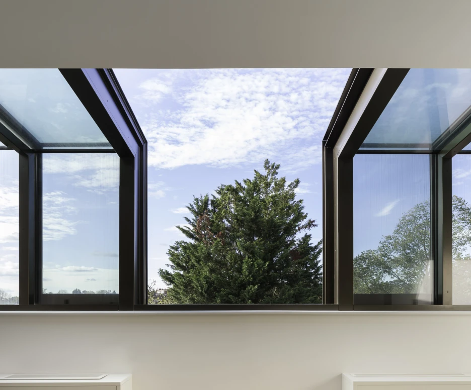 Ventana amplia con marcos oscuros, mostrando un paisaje exterior con un árbol y cielo parcialmente nublado. Interior moderno con pared blanca y suelos de madera clara.