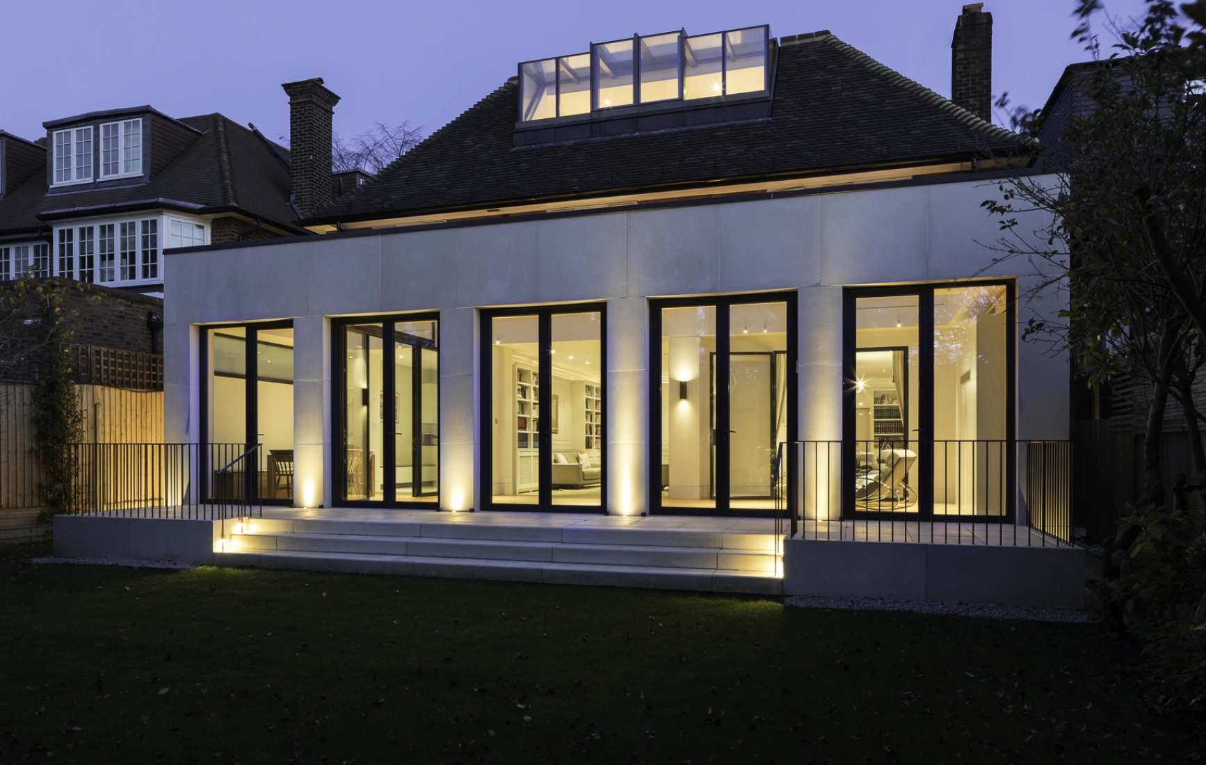 Casa moderna al anochecer, con fachada de grandes ventanales que iluminan el interior. El jardín está bien cuidado y la iluminación exterior resalta la arquitectura.