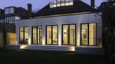 Maison moderne avec de grandes fenêtres illuminées au crépuscule. Le jardin est soigné, entouré de clôtures en bois. Un toit en pente et un vitrage au-dessus de la porte ajoutent du caractère.