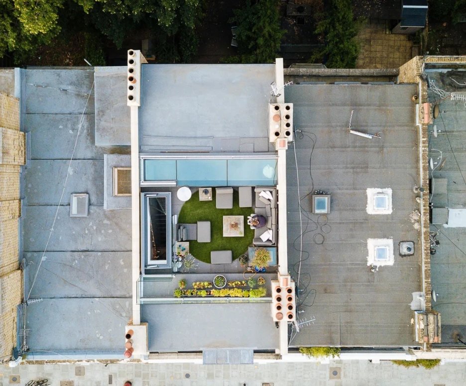 Aerial view of a modern rooftop garden featuring green grass, outdoor seating, and planters. Surrounding buildings and trees are visible, along with parked cars on the street below.