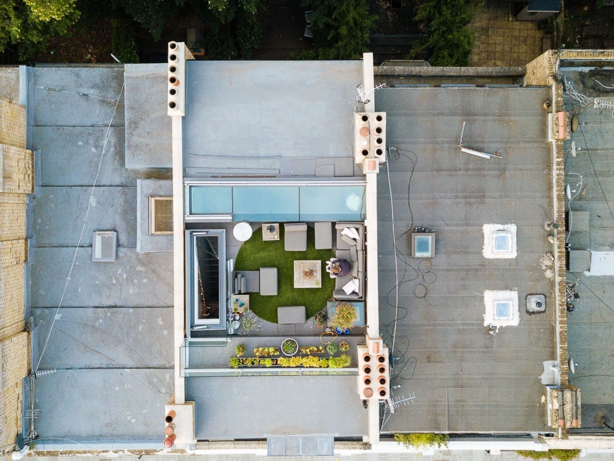 Aerial view of a modern rooftop terrace featuring a green lawn, seating area with outdoor furniture, and planters. Surrounding buildings and a tree-lined street are visible below.