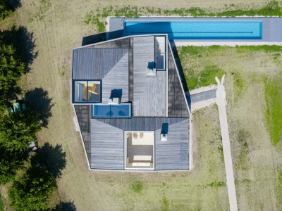 Vue aérienne d’une maison moderne aux lignes angulaires, entourée de verdure. Une piscine s’étend le long de la propriété, avec une allée menant à l’entrée.