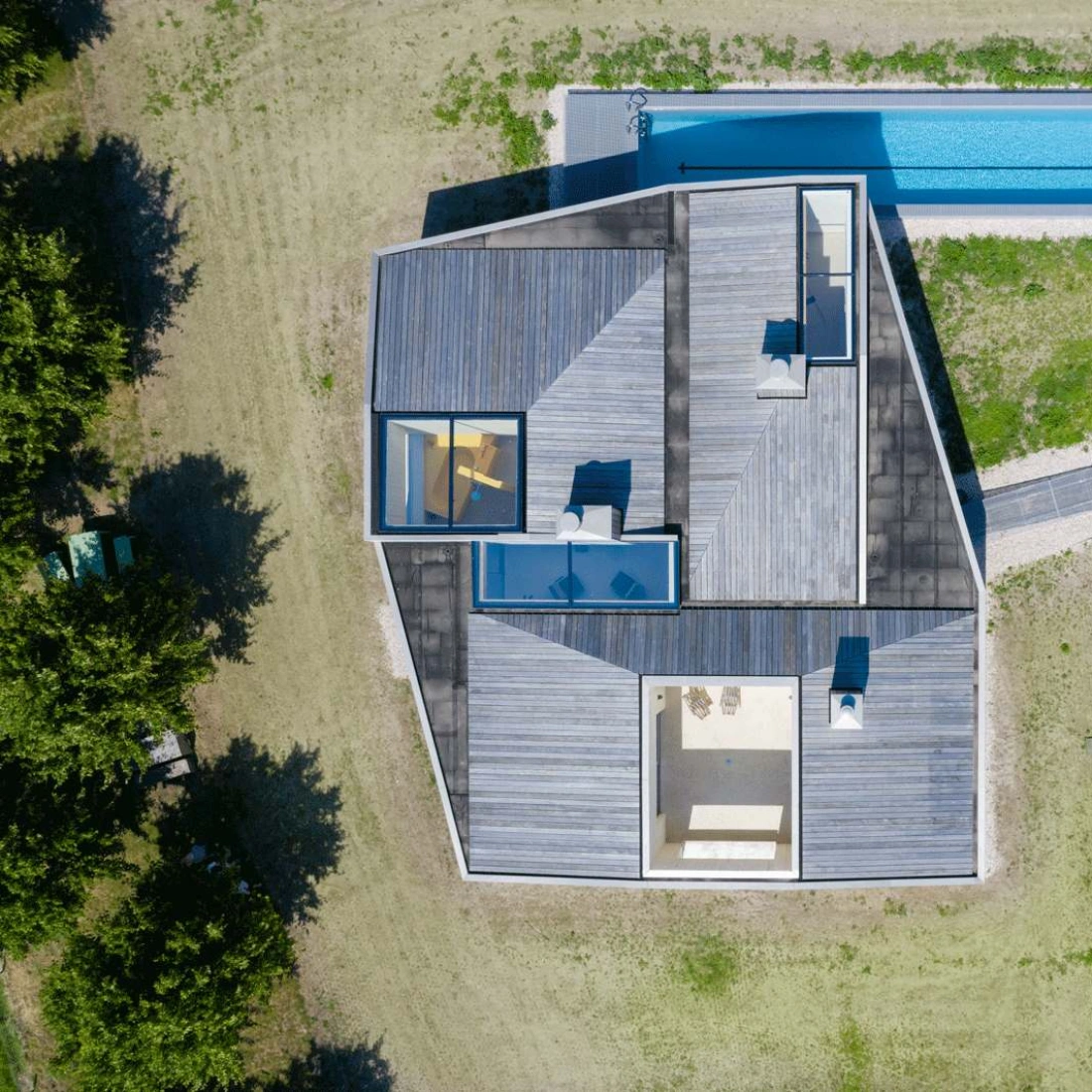 Aerial view of a modern, angular house featuring a flat roof and wooden deck. A rectangular swimming pool runs alongside the house, surrounded by a grassy lawn.