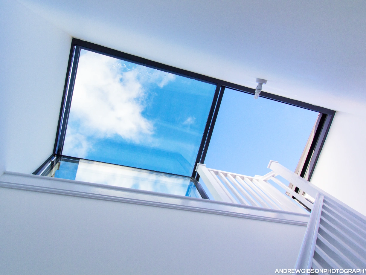 A view looking up through a large skylight, revealing a bright blue sky with a few fluffy clouds. The interior features white walls and a staircase leading to the skylight.