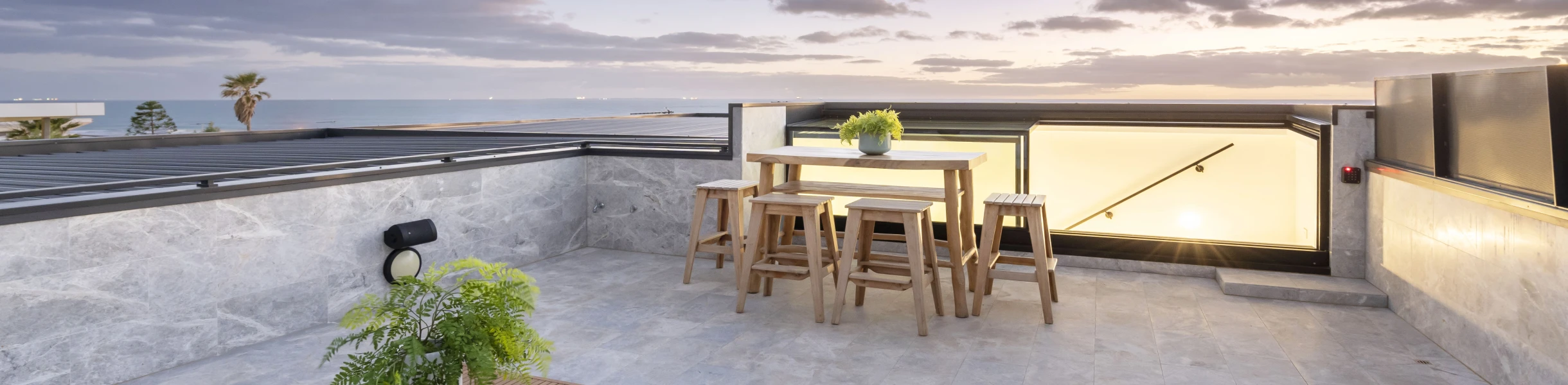 A modern rooftop terrace features a wooden table surrounded by high stools, with a stylish coffee table and lush greenery. The scene is illuminated by twilight, showcasing a serene ocean view and scattered clouds in the sky.