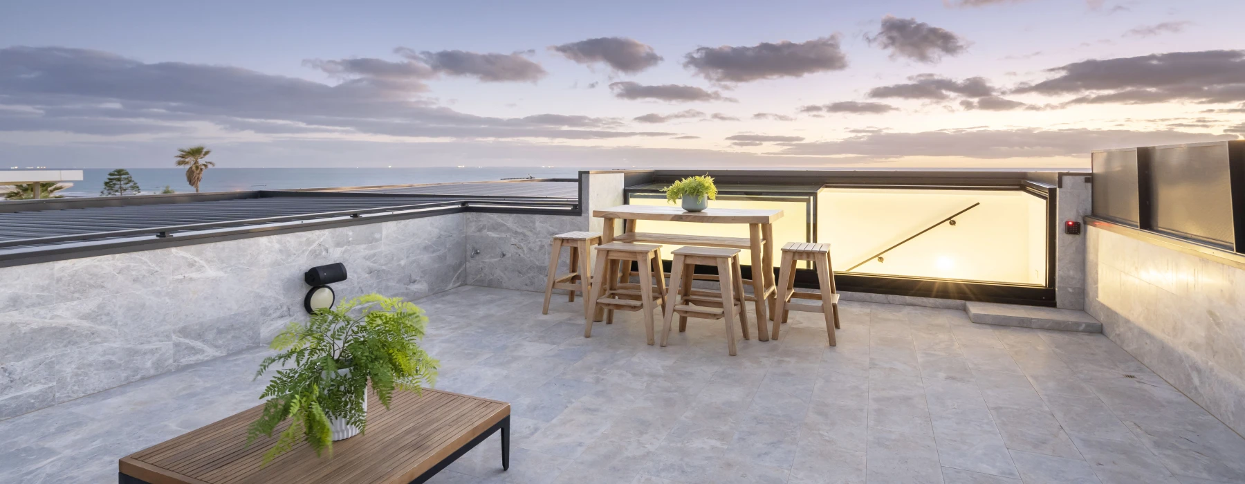 A modern rooftop terrace features a wooden table surrounded by high stools, with a stylish coffee table and lush greenery. The scene is illuminated by twilight, showcasing a serene ocean view and scattered clouds in the sky.