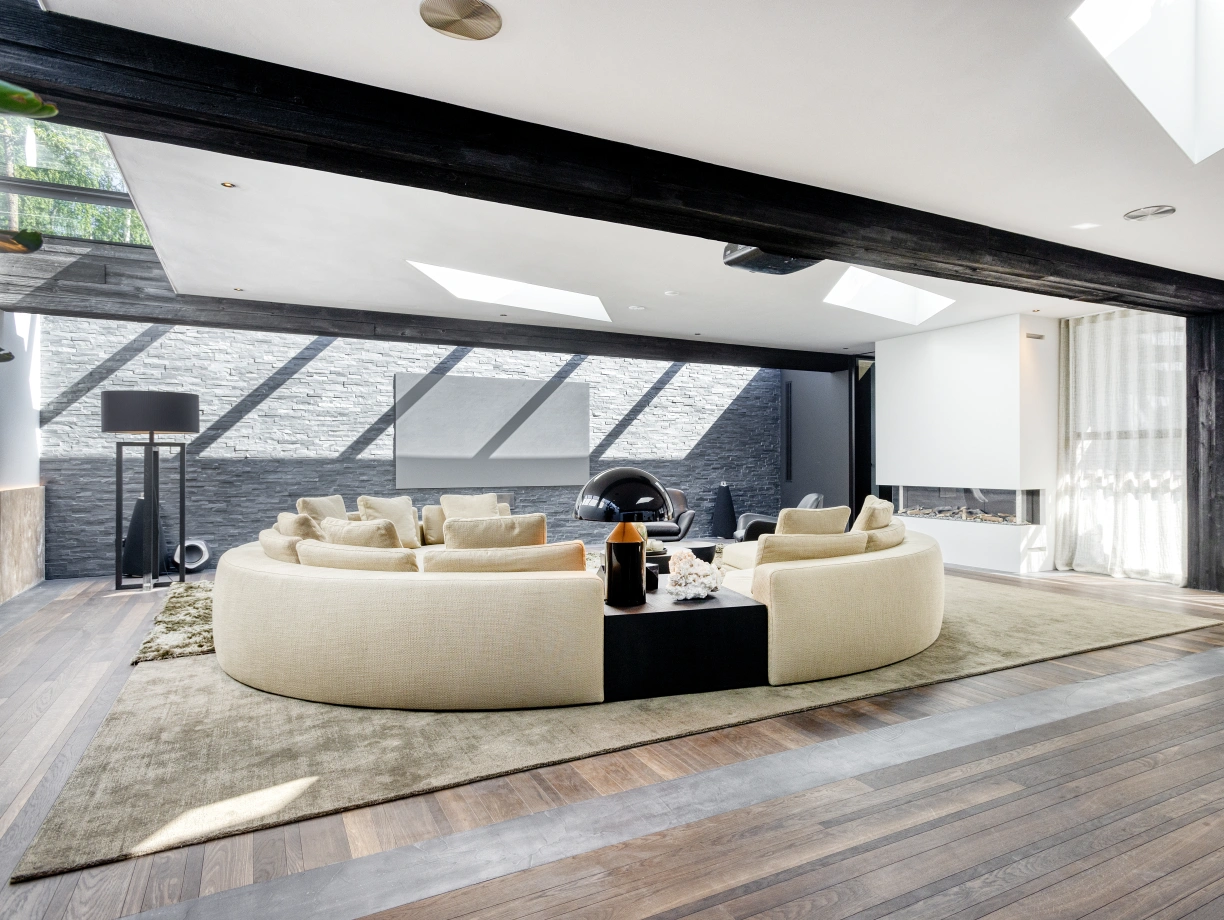 Sala moderna con un diseño abierto. Sofás en forma de curva rodean una mesa baja negra. Ventanas grandes permiten la entrada de luz natural. Suelo de madera clara y paredes de piedra decoran el espacio.