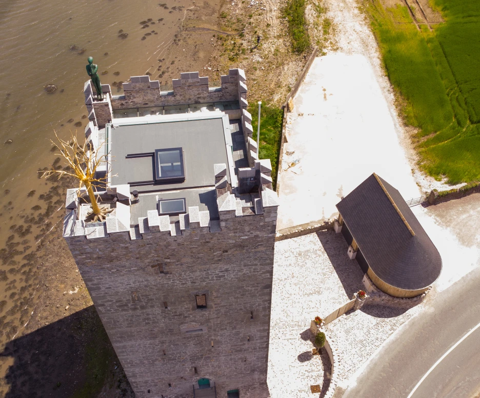 Vista aérea de un castillo de piedra, con un techo plano y una estatua en la parte superior. Al lado, hay una construcción moderna y un camino que rodea la edificación. El entorno es natural, con un área de agua cercana y campos verdes.
