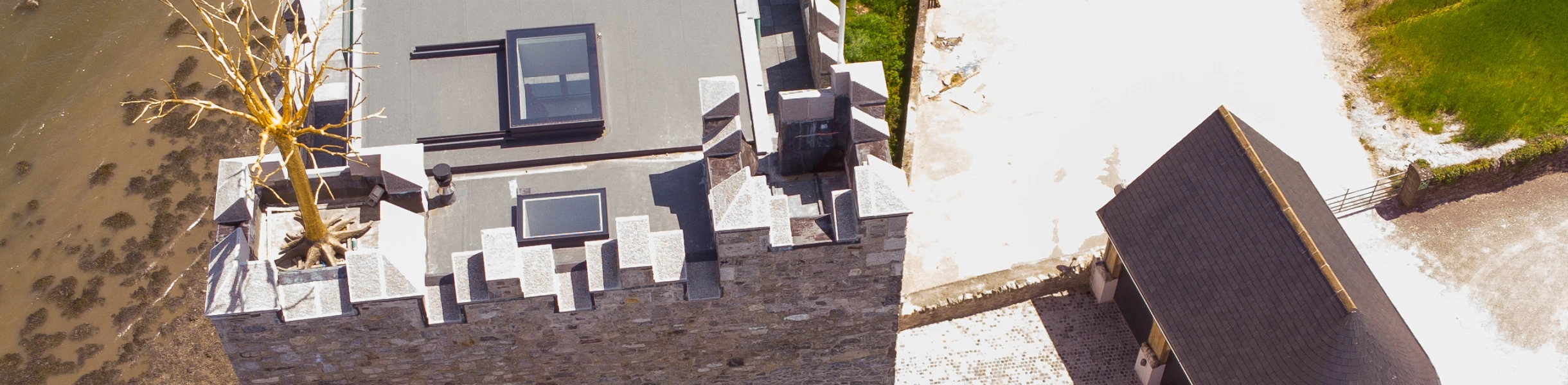 Aerial view of a historic stone tower situated near a riverbank, featuring a modern rooftop terrace and a nearby circular building. The surroundings include a gravel pathway and lush greenery.