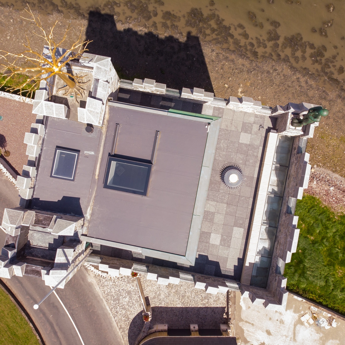 An aerial view of a unique castle-like structure positioned near a shoreline. The roof is flat with skylights, and there is a decorative element resembling a tree. Surrounding the building are stone walls and a road, with a grassy area visible.