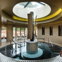 A serene spa interior featuring a circular hot tub with elegant water features, surrounded by mosaic tiles. Natural light streams in through a skylight, illuminating the space, while plush towels are neatly arranged nearby.