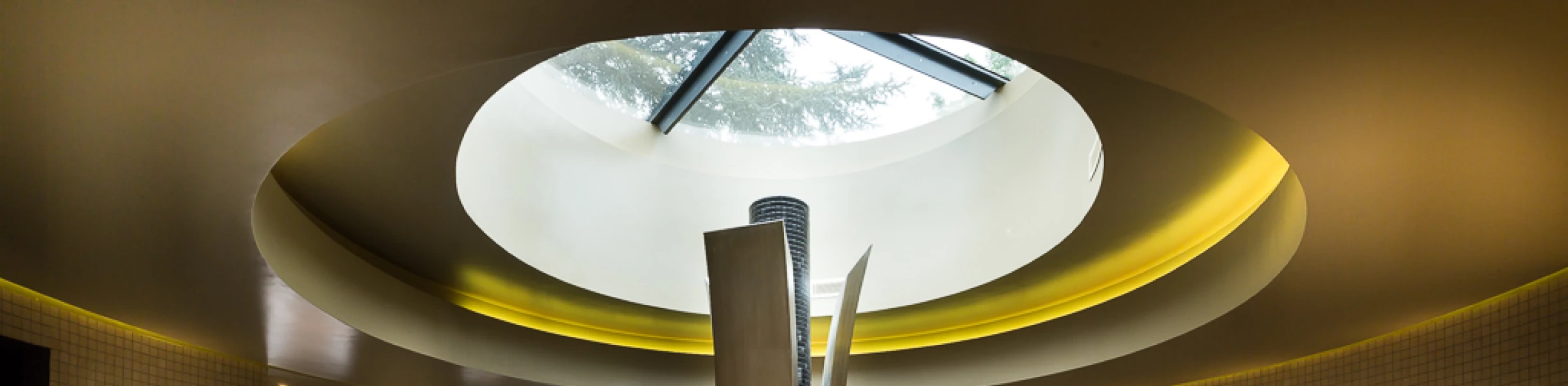 Spa moderno con una fuente central y jacuzzi. La iluminación suave resalta las paredes de azulejos claros y las ventanas permiten la entrada de luz natural. Toallas enrolladas y un ambiente relajante completan la escena.