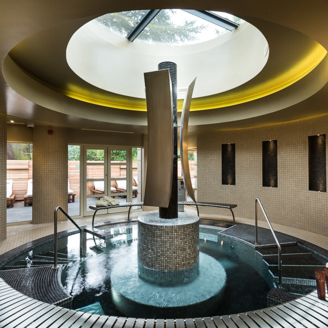 A serene spa interior featuring a circular hot tub with elegant water features, surrounded by mosaic tiles. Natural light streams in through a skylight, illuminating the space, while plush towels are neatly arranged nearby.
