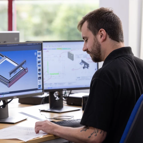 A man sitting at a desk, focusing on a computer screen displaying a 3D model and engineering designs. He has a notepad and documents in front of him. Two additional monitors are visible in the background.