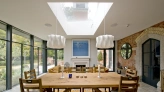 A modern dining room features a large wooden table surrounded by chairs. Two stylish white pendant lights hang above the table, and a large skylight allows natural light to fill the space. A fireplace with a simple design contrasts with the brick wall. Large glass doors offer a view of the outdoor area.