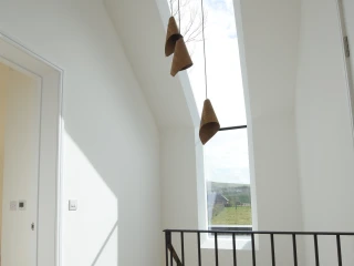 Un couloir lumineux avec un plafond en pente et une grande fenêtre ouverte sur le ciel. Trois suspensions en forme de cône sont accrochées au plafond. Un escalier en bois est visible à gauche, avec une balustrade noire minimaliste.