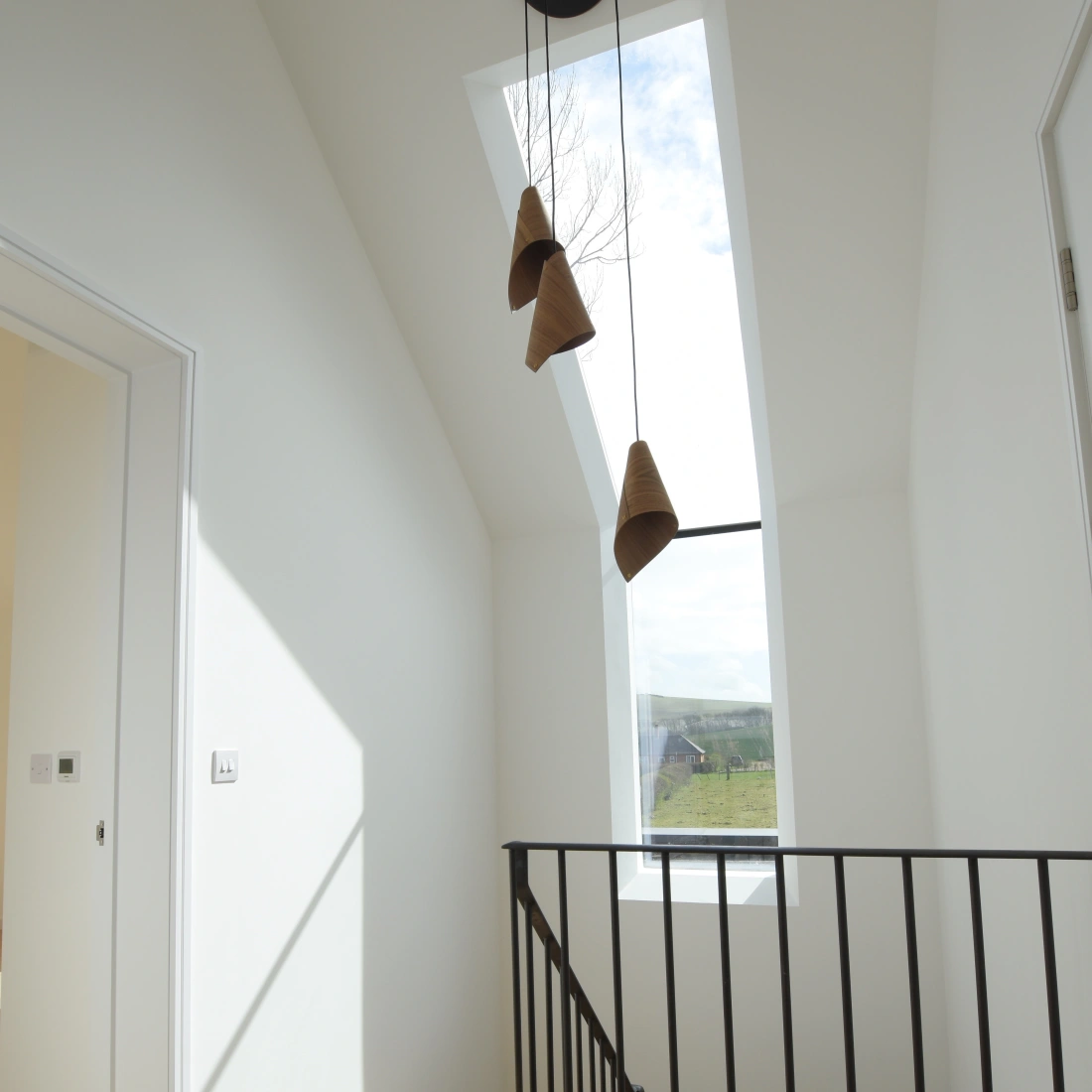 Pasillo iluminado con pared blanca y escalera de madera. Ventana de techo que permite la entrada de luz natural. Tres lámparas colgantes de forma cónica.